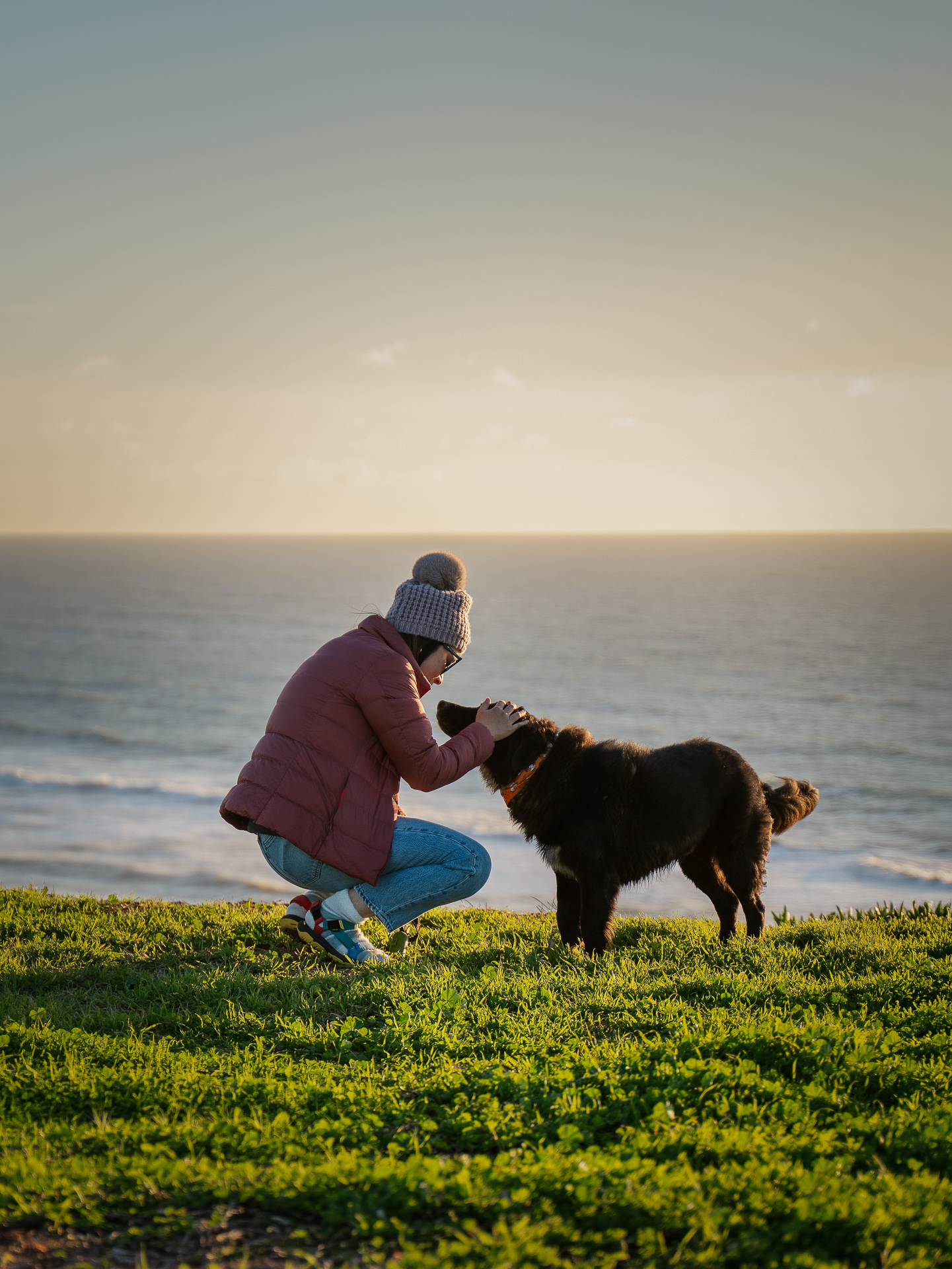 Woman kneeling to pet her dog on a grassy coastal cliff at sunset
